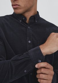 Navy corduroy shirt with a button-down collar, front pocket, and buttoned cuffs. Features textured stripes and contrasting buttons.