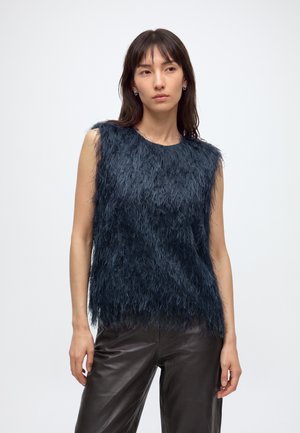 Femme portant un haut sans manches bleu foncé à texture duveteuse et un pantalon en cuir noir, debout devant un fond clair uni.