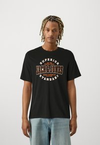 Black t-shirt with short sleeves, featuring a bold, vintage-style orange graphic text "JACK & JONES" centered on the front. Casual design.