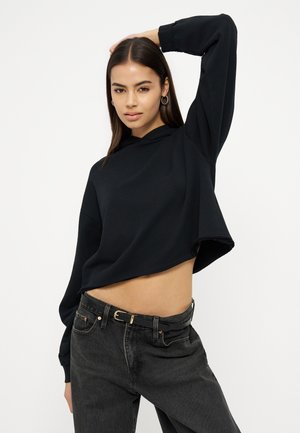 Woman with long dark hair wearing black cropped hoodie and high-waisted black jeans, raising one arm with a neutral expression.