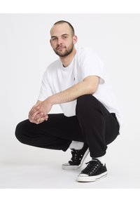 T-shirt blanc, pantalon de survêtement noir et baskets noires avec semelles blanches. Le modèle est accroupi sur une surface plane, les mains jointes, avec une expression neutre.