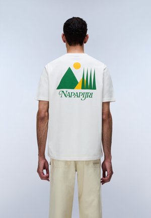 Man facing away wearing white T-shirt with green and yellow mountain, trees graphic and "NAPAPIJRI" text, paired with light beige pants.
