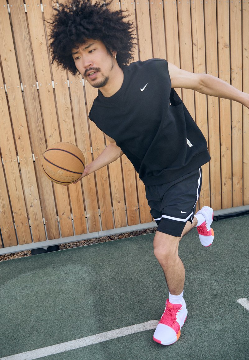 Maillot sans manches noir et shorts noirs avec des accents blancs ; baskets roses et blanches. Un joueur dribble un ballon de basket sur un terrain.