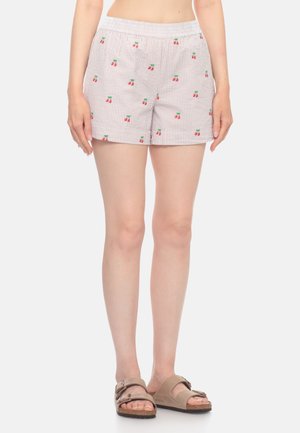Persona che indossa shorts chiari a righe con piccole stampe di ciliegie rosse e sandali beige, in piedi su uno sfondo bianco semplice.
