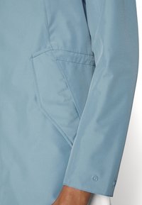 Helle blaue Jackenärmel mit aufgenähtem Taschen und Druckknopfmanschette, getragen von einer Person mit sichtbarem, dunklem Hautton der Hand.
