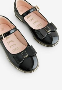 Zapatillas de charol negro estilo Mary Jane con punta redonda, ribete dorado y un lazo decorativo con herrajes dorados en la correilla frontal.
