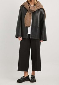 Zwarte leren jas, oversized pasvorm, ritssluiting, gecombineerd met een witte t-shirt en wijde zwarte broek. Bruine gebreide sjaal om de nek gewikkeld.