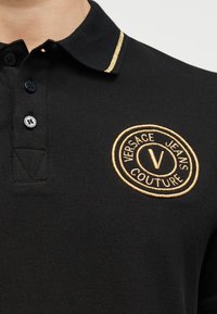 Μαύρο πόλο πουκάμισο με χρυστό κεντημένο λογότυπο Versace Jeans Couture στο στήθος και χρυσή επένδυση στον γιακά.