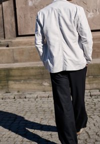 Mężczyzna idący po brukowanej nawierzchni w pobliżu kamiennego muru, ubrany w białą kurtkę i czarne spodnie, z rękami w kieszeniach.