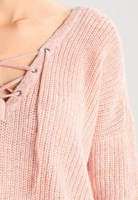 Maglione a maglia rosa chiaro con scollatura a V, caratterizzato da lacci incrociati e occhielli metallici. La texture è morbida e a costine, con spalle scese.