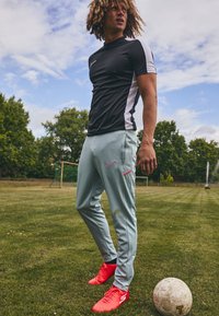 Svart og hvit idrettsdrakt, lyseblå bukser med rosa detaljer, og røde fotballsko. Står på gresset med en fotball.
