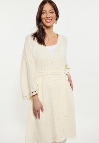 Lichtbeige lange cover-up met scalloped rand, wijde mouwen, strikriem en textuurstof. Draag het over een witte top.