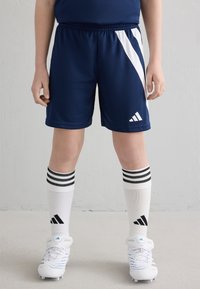 Sinine sportlikud lühikesed püksid valgete külgpindadega, musta ja valge triibuga sokid ning valged jalgpallijalad siniste aktsentidega. Adidas brändingu kohal.