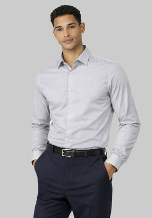 Homme aux cheveux courts et bouclés portant une chemise gris clair à boutons et un pantalon bleu marine foncé avec une ceinture noire, debout les mains dans les poches.