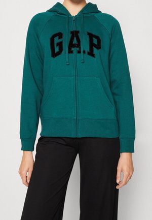 Person iført grøn lynlåstrøje med sorte "GAP" bogstaver foran og sorte bukser, vist fra nakke til hofter mod en ensfarvet baggrund.