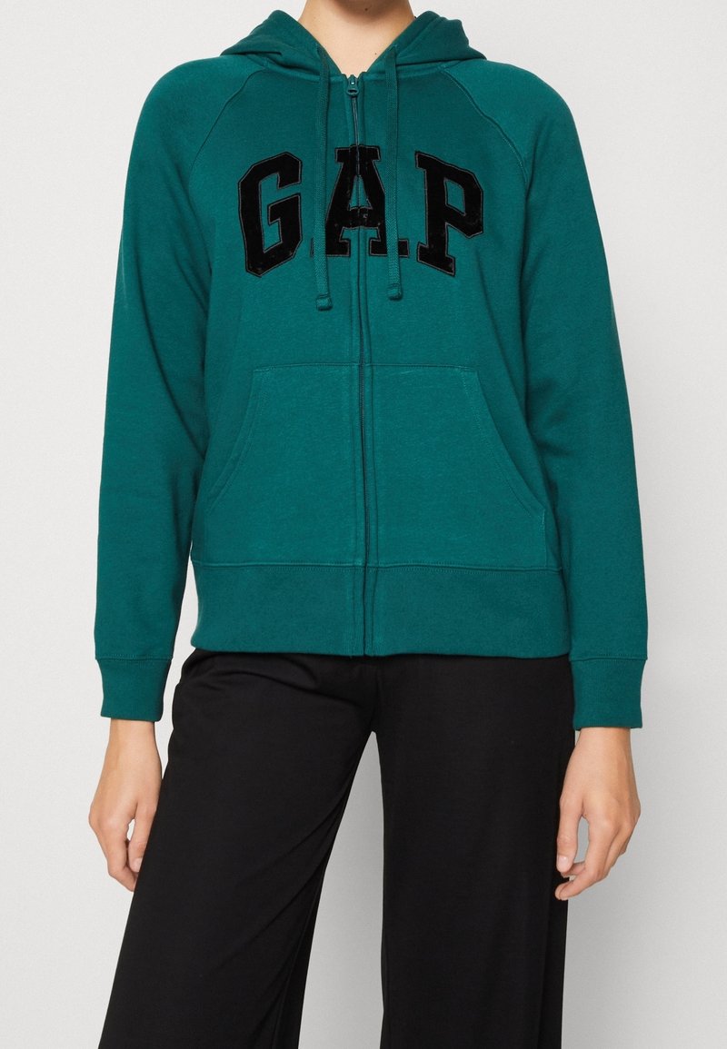 Personne portant un sweat à capuche zippé vert avec des lettres noires "GAP" sur le devant et un pantalon noir, visible du cou jusqu’aux hanches sur un fond uni.