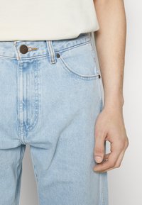 La mano derecha descansando junto a unos jeans de mezclilla azul claro, combinados con una blusa blanca metida por dentro, mostrando detalles del botón y el bolsillo de los jeans.