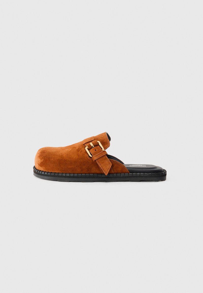 Bruna mocka slip-on klackar med svart sula och justerbart guldbuckle-band mot en enkel ljus bakgrund.