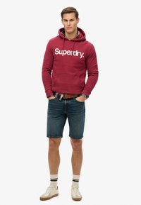 Red hoodie with white "Superdry" logo, blue denim shorts, white sneakers with rubber soles, and cream striped socks. Casual outfit.