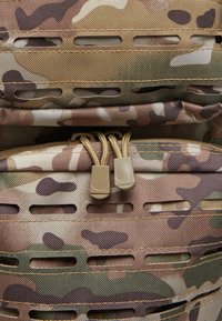 Camouflage backpack material features beige, brown, and green patterns with horizontal slot designs and golden zipper pulls. Textured finish.
