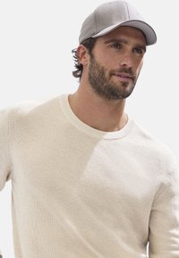 Beige gestrickter Pullover mit Rundhalsausschnitt, der ein strukturiertes Muster aufweist. Graue Baseballmütze auf einem Mann mit welligem Haar und Bart.