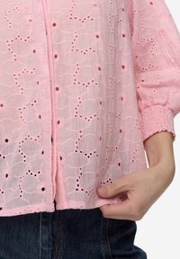 Lichtroze blouse met geborduurde bloemenpatronen met ajourdetails, pofmouwen en gerimpelde manchetten. De stof is lichtgewicht en heeft een textuur.