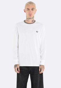 Maglietta a maniche lunghe bianca con strisce nere sul colletto, con un piccolo emblema nero sul petto. Tessuto morbido, vestibilità casual.