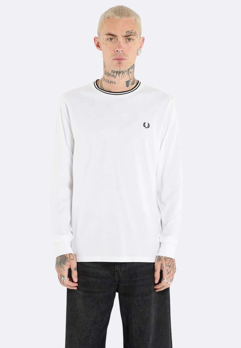 Maglietta a maniche lunghe bianca con strisce nere sul colletto, con un piccolo emblema nero sul petto. Tessuto morbido, vestibilità casual.