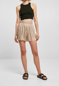 Top court noir à côtes, short en satin beige avec taille élastique, et sandales noires à enfiler, se tenant sur un fond neutre.