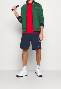 Man i grön zip-up jacka, röd skjorta, marinblå shorts och vita sneakers som håller en tennisracket över axeln mot en neutral bakgrund.
