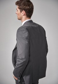 Veste de costume grise sur mesure avec un tissu texturé, revers standard et deux boutons. La vue de dos montre des épaules structurées et une silhouette élégante.