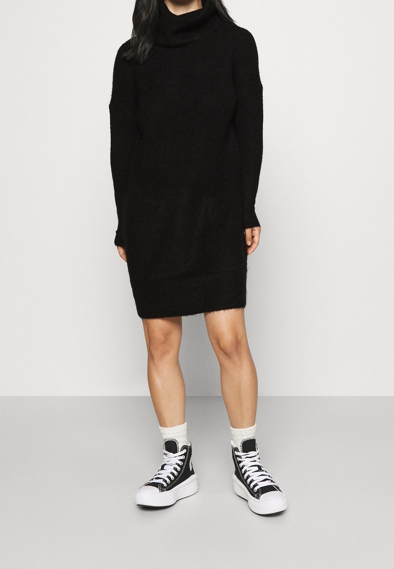 Personne portant une robe-pull à col roulé noire à manches longues et des baskets montantes noires avec des semelles et des lacets blancs.