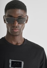 Gafas de sol angulares transparentes con lentes oscuros, llevadas sobre una camiseta negra con un logotipo gráfico en blanco y gris. Primer plano, fondo neutro.