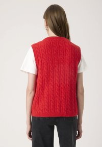 Woman with long brown hair wearing silver hoop earrings, a bright red cable-knit sleeveless sweater over a white short-sleeve shirt, and black jeans.