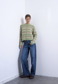 Γυναίκα ακουμπισμένη σε λευκό τοίχο, φοράει πράσινη ριγέ ζακέτα με κουμπιά, τζιν παντελόνι με φαρδιά μπάτα και σκούρα καφέ παπούτσια.