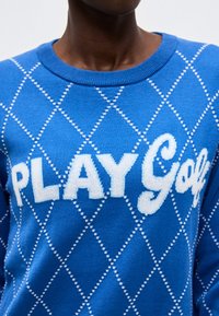 Μπλε πουλόβερ με λευκό διαμαντέ μοτίβο και μεγάλο λευκό κείμενο "PLAY Golf" στο στήθος ενός ατόμου με σκουρόχρωμο δέρμα.