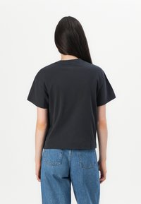 Donna con capelli lunghi e scuri che indossa una semplice maglietta nera a maniche corte e jeans blu, in piedi di spalle contro uno sfondo bianco.