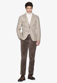 Beige hørfiskeblazer med spidse klipper, hvid turtleneck og brune fløjl bukser. Brune snørebåndssko fuldender outfittet.