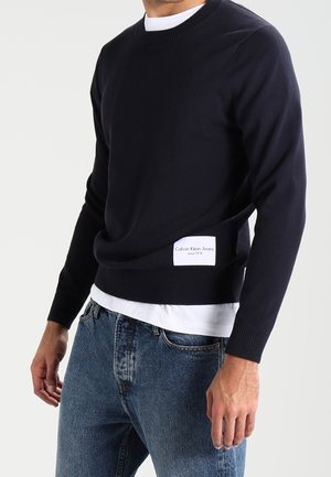 Mann, der einen marineblauen Calvin Klein Jeans Pullover über einem weißen Hemd mit blauen Jeans trägt, vor einem schlichten Hintergrund stehend.