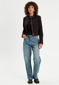 Black cropped jacket with a front zip and stand collar, paired with light blue straight-leg jeans and black chunky boots. Curly hair and earrings.