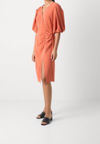 Robe orange rouille avec manches bouffantes, décolleté en V, fronces sur le côté et une fente haute sur la cuisse ; portée avec des sandales à talons ouvertes noires.