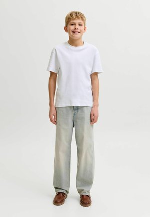 Šypsantis berniukas su trumpais šviesiais plaukais, dėvintis paprastą baltą marškinėlį, laisvus šviesiai mėlynus džinsus ir rudus batus, stovintis prie vienarūšio šviesaus fono.