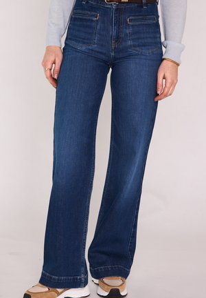 Personne portant un jean flare en denim bleu foncé, des baskets beige et blanches, un haut à manches longues gris clair, et une ceinture marron, debout devant un fond neutre.