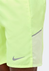 Shorts de sport jaune vif avec panneau latéral perforé et logo Nike swoosh gris au-dessus du genou sur une jambe à la peau foncée.