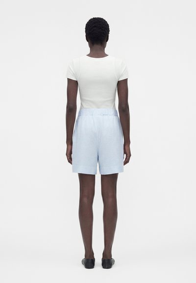 Άτομο που στέκεται με την πλάτη προς τα εμπρός, φοράει λευκό κοντομάνικο πουκάμισο, ανοιχτό μπλε υφασμάτινες βερμούδες και μαύρα παπούτσια χωρίς κορδόνια.