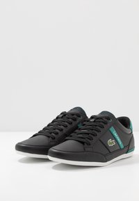 Mustanväriset matalat tennarit, joissa sileä nahkapinta, turkoosit yksityiskohdat ja valkoinen kumipohja. Mallissa on nauhakiinnitys ja logo.