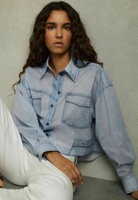 Camicia di jeans oversize azzurro chiaro con due tasche frontali, bottoni neri e colletto. Tessuto morbido, design corto.