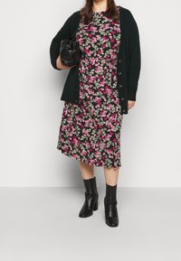 Schwarzes Blumenkleid mit rosa und grünen Mustern, darunter eine dunkelgrüne Strickjacke. Schwarze Stiefeletten mit Absatz und eine schwarze Handtasche.