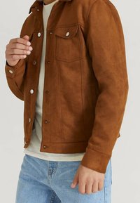Chaqueta de ante marrón con cuello, bolsillos frontales y botones a presión. Llevada sobre una camiseta de color beige claro, combinada con jeans azules.