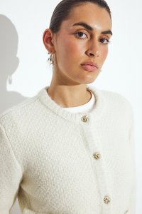 Balta tekstūru kardiganas su apvaliu iškirpimu, puošiamas dekoratyviniais aukso spalvos mazgų mygtukais ir įribuoti apykaklės detalėmis.